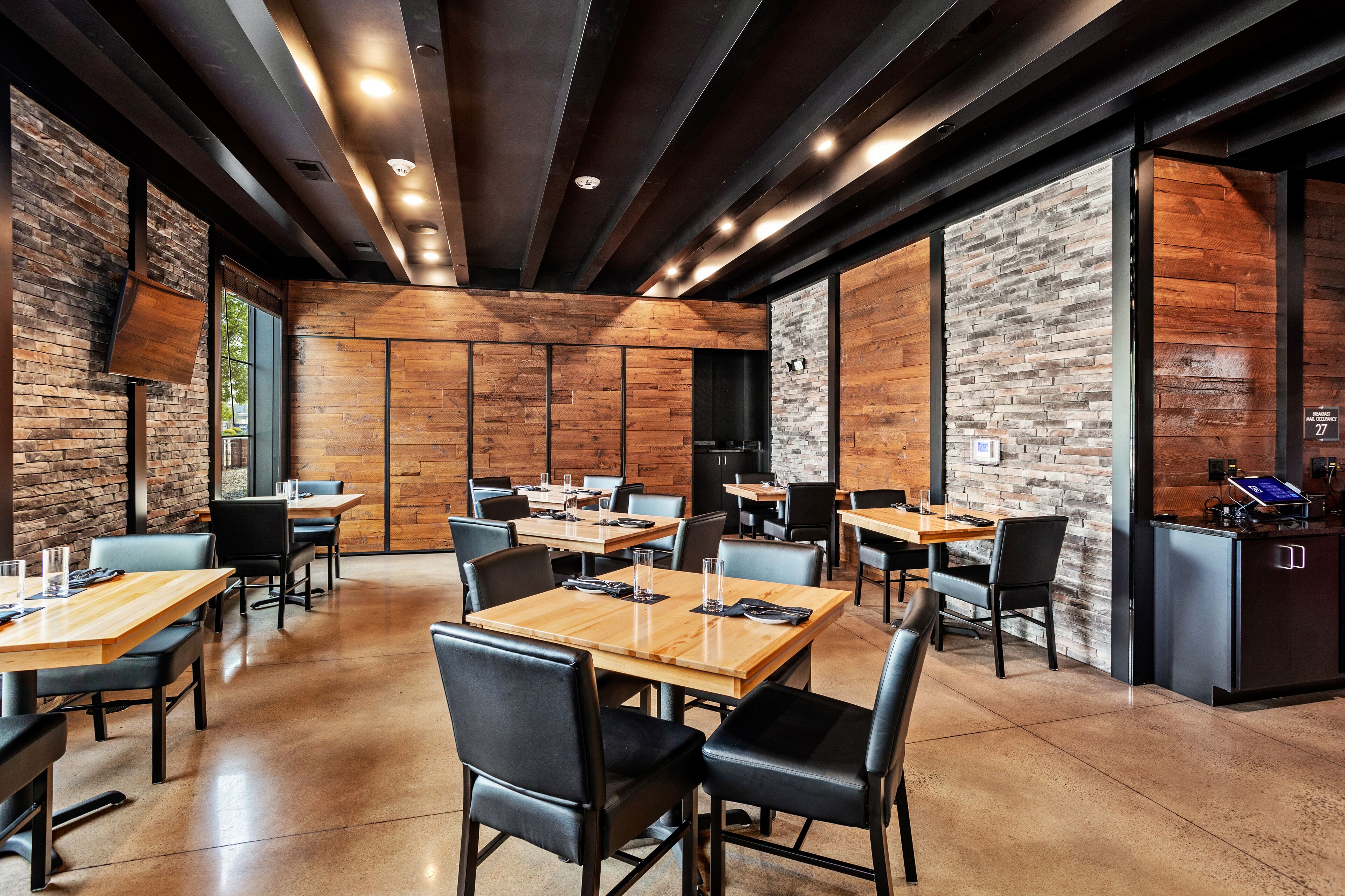 A modern dining area with wooden tables and black chairs, featuring a stone wall and a black cabinet with a digital screen.
