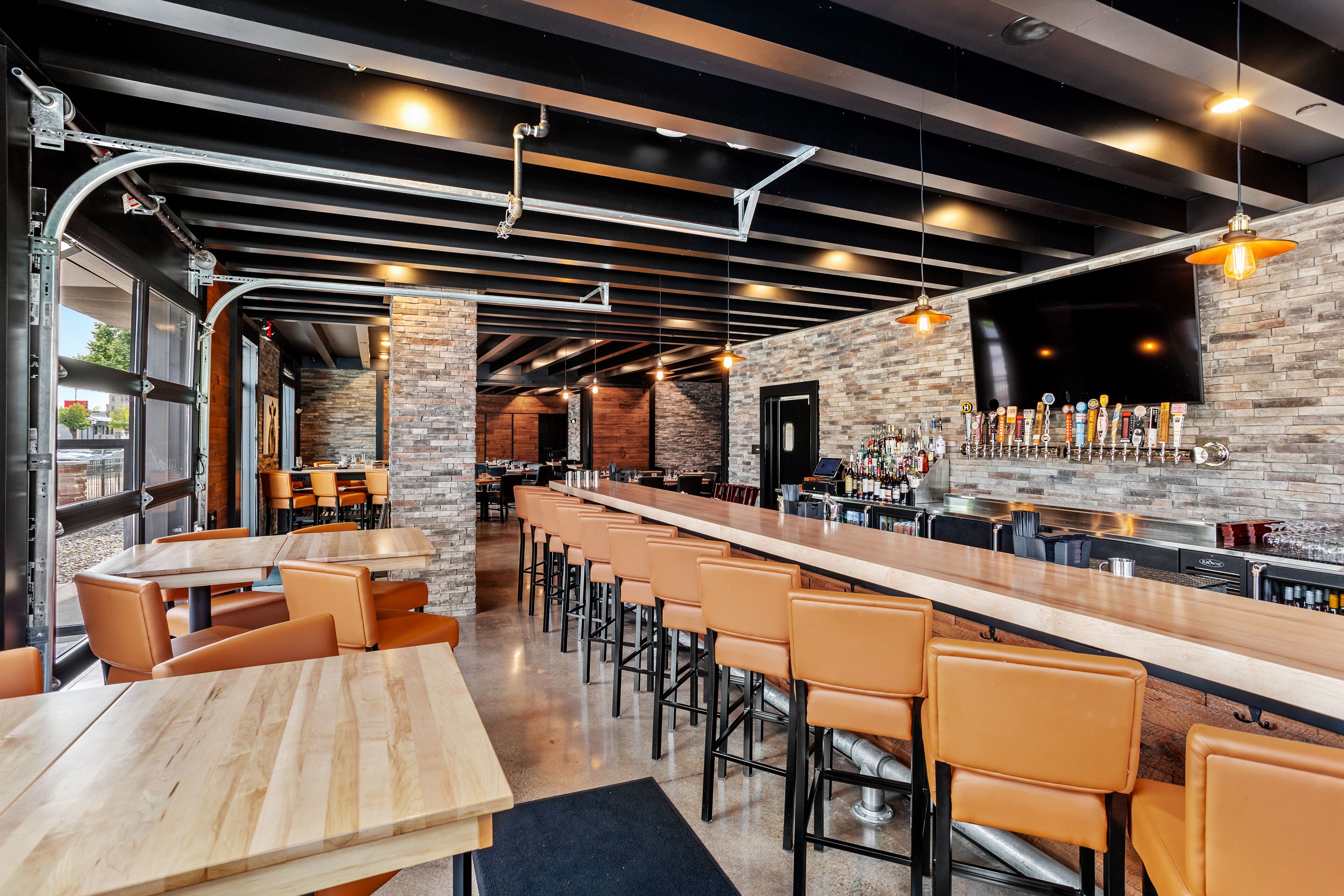 A modern bar area with a long counter, multiple stools, and a brick wall backdrop.