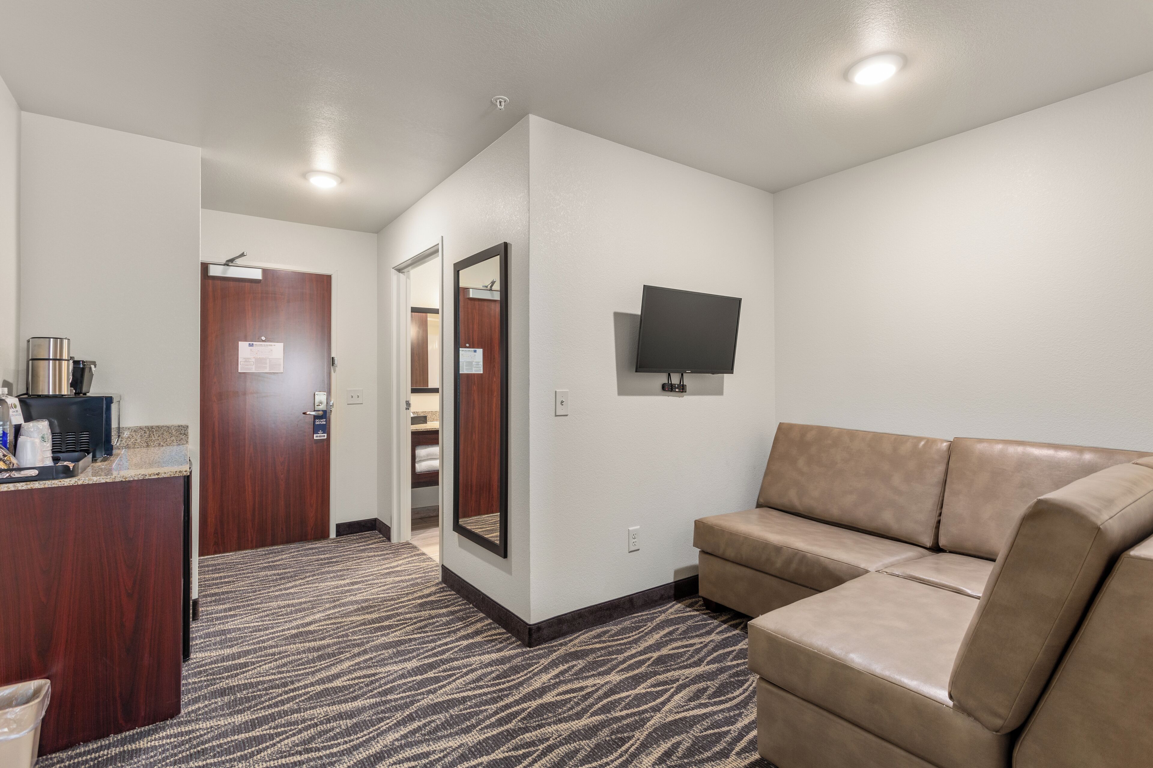 A hotel room with a kitchenette, a large mirror, a flat-screen TV, and a beige sofa.