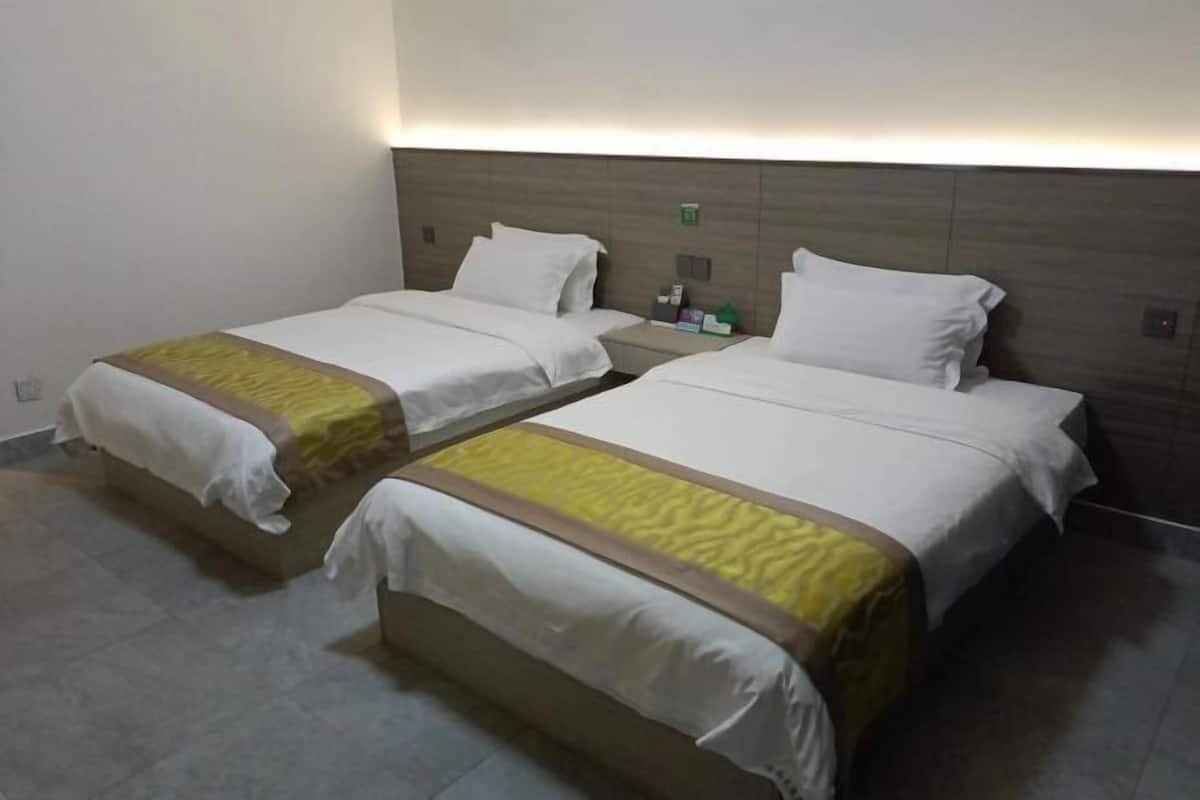 Dos camas individuales con ropa de cama blanca y detalles amarillos, una mesita de noche con artículos de aseo, y una lámpara fijada a la pared.