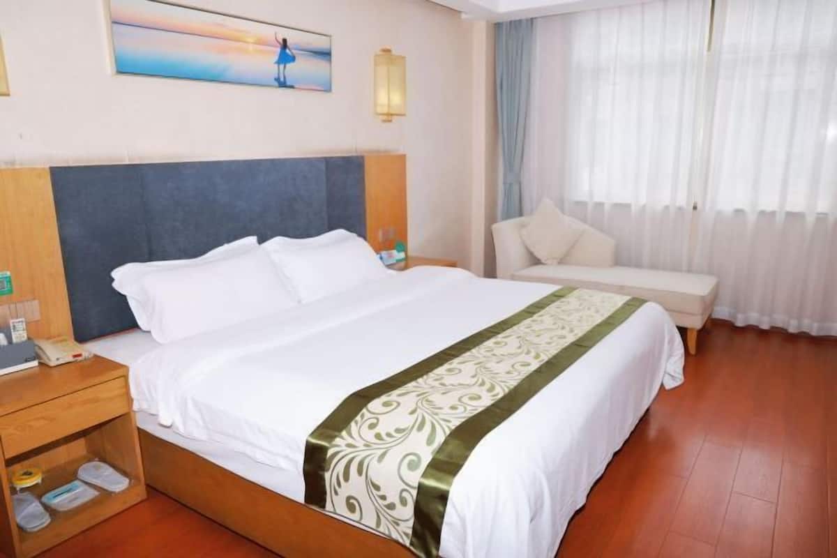 Una habitación de hotel con cama, mesita de noche, silla, un cuadro en la pared y ventana con cortinas.