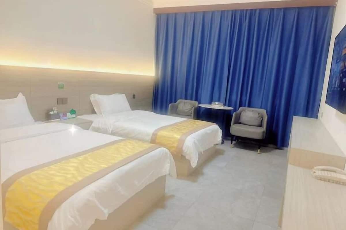 Habitación de hotel con dos camas, una mesita pequeña, dos sillas y un amplio ventanal con cortinas azules.