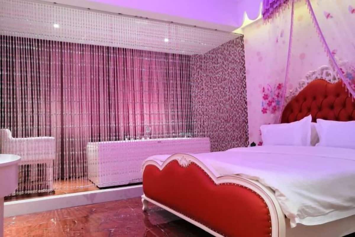 Un dormitorio con cama tapizada en rojo, ropa de cama blanca y un cabecero decorativo. En la esquina, hay una silla blanca y una mesita.