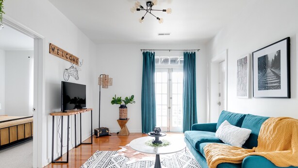 Una sala de estar moderna con un sofá color turquesa, una mesa de centro y un televisor sobre un mueble. Hay un cartel con la palabra 'WELCOME' en la pared y un cuadro enmarcado sobre el sofá.
