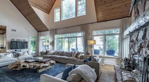 A spacious living room with a high ceiling, large windows, a stone fireplace, and a cozy seating area.