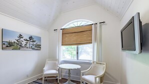 A room with a white ceiling, a painting of trees, a window with a bamboo blind, two white chairs, a small round table, and a TV mounted on the wall.
