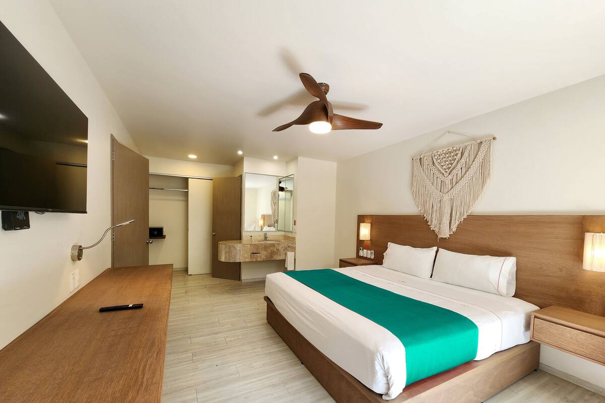 Ein Hotelzimmer mit Bett, Nachttisch, Fernseher, Deckenventilator und einem im Hintergrund sichtbaren Badezimmer.
