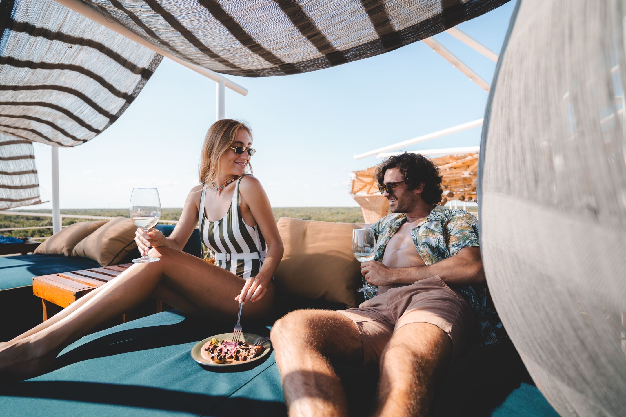 A couple relaxing on a sun lounger with a view of the ocean, a plate of food, and drinks.