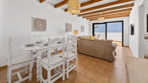Un comedor con mesa y sillas blancas, un sofá y vistas al mar a través de un ventanal.