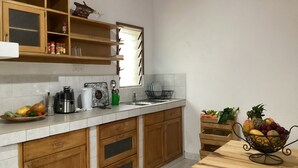 A kitchen with wooden cabinets, a white countertop, a coffee maker, a fruit bowl, and a window with blinds. - Villa ChezSoa Andoharanofotsy (ANTANANARIVO)