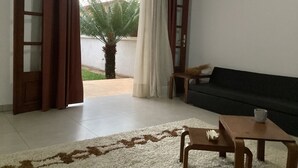 A living room with a sofa, a coffee table, and a chair. There is a window with curtains, a door, and a view of a palm tree outside. - Villa ChezSoa Andoharanofotsy (ANTANANARIVO)