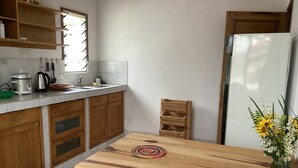 A kitchen with wooden cabinets, a wooden dining table, a refrigerator, and a window with blinds. - Villa ChezSoa Andoharanofotsy (ANTANANARIVO)