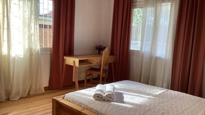 A bedroom with a wooden bed, a wooden desk, and a chair. There is a window with curtains, and a small table with a vase of flowers. - Villa ChezSoa Andoharanofotsy (ANTANANARIVO)