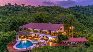Un resort de dos pisos con piscina, mobiliario exterior y vistas al bosque circundante.