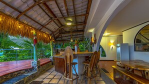 Un espacio exterior techado con barra de madera, asientos y vistas a la vegetación.