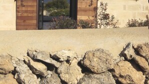 Un edificio de piedra con puerta y contraventanas de madera, un muro de piedra en primer plano, y vista al mar a través de la puerta.