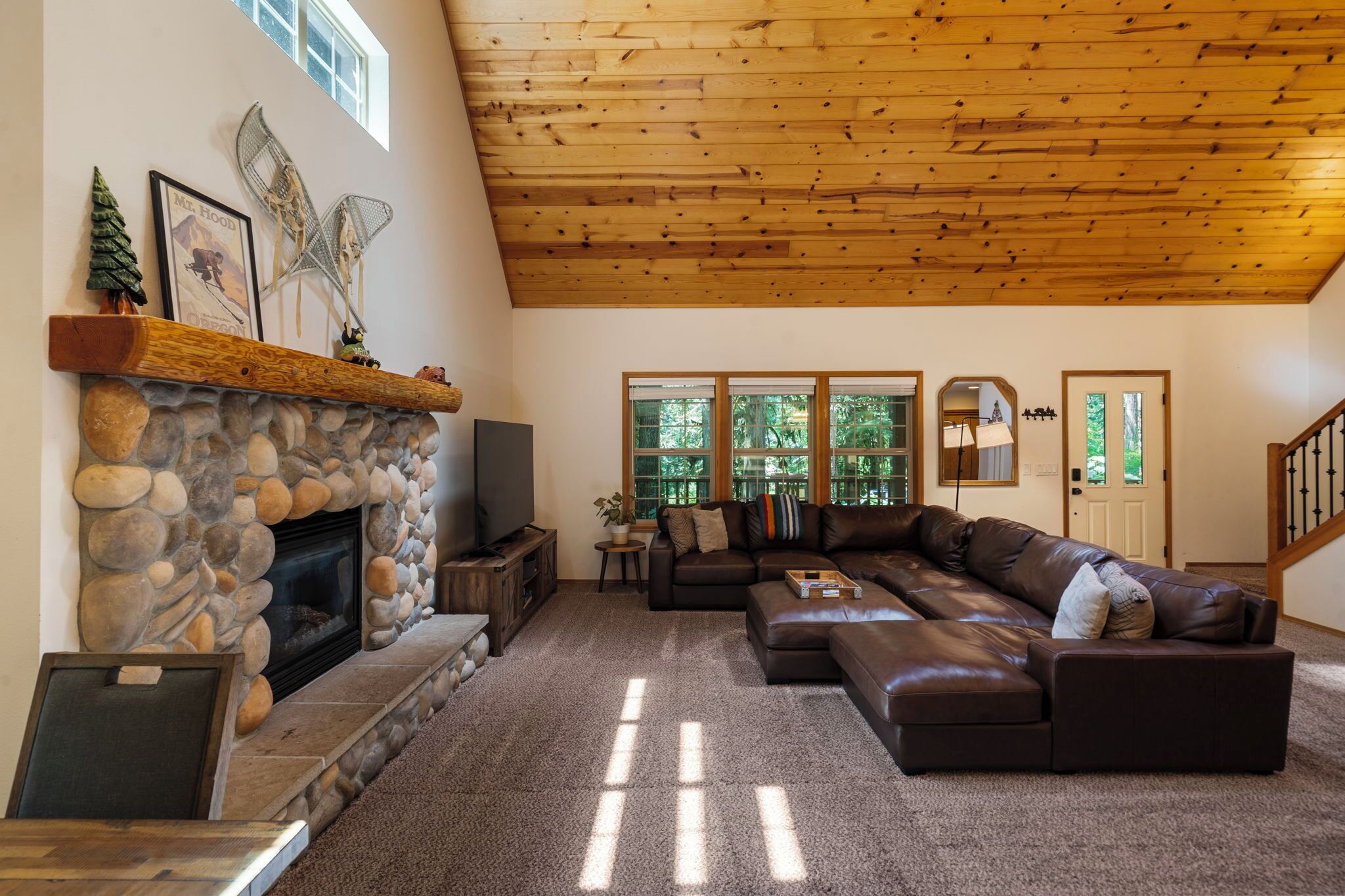 A living room with a stone fireplace, leather sofa, wooden ceiling, and large windows.