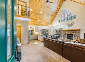 A spacious living area with a high ceiling, a fireplace, and a kitchen in the background. - Cozy Cascade Cabin. Relaxing Hot Tub, Spacious and Creek Nearby (Rhododendron)
