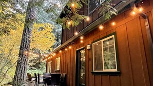 A wooden cabin with a deck, string lights, and a dining area with chairs and a table. - Cozy Cascade Cabin. Relaxing Hot Tub, Secluded, Spacious and Creek Nearby (Rhododendron)