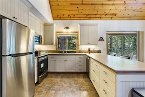 A modern kitchen with a stainless steel refrigerator, built-in oven, and microwave. It features light-colored cabinetry, a large island with a sink, and a wooden ceiling.