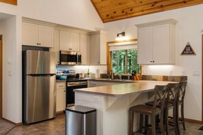 A modern kitchen with stainless steel appliances, a large island with bar stools, and a view of the outdoors through a window. - Cozy Cascade Cabin. Relaxing Hot Tub, Spacious and Creek Nearby (Rhododendron)
