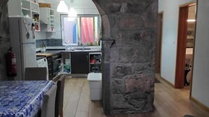 A dining area with a table and chairs, a kitchen with a stone archway, and a living room with a sofa and a patterned pillow. - House (Ribeira Grande)