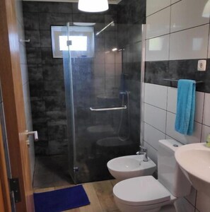 A bathroom with a glass shower enclosure, two toilets, a sink, and a blue towel on the wall.
