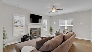 A modern living room with a brown sofa, a fireplace, a ceiling fan, and large windows. - Spacious 4 BD Golf Getaway with Game Room! (Aberdeen)