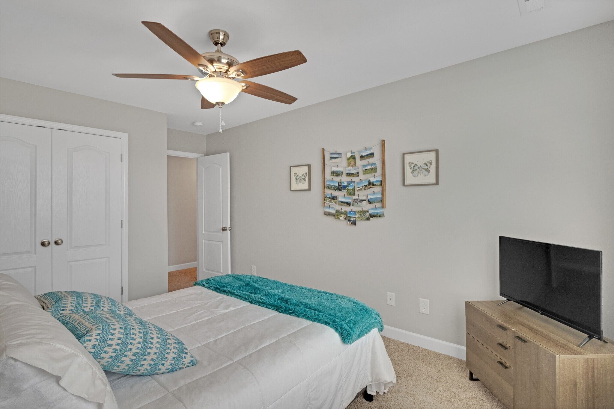 A bedroom with a bed, a ceiling fan, a TV on a wooden dresser, and framed pictures on the wall.