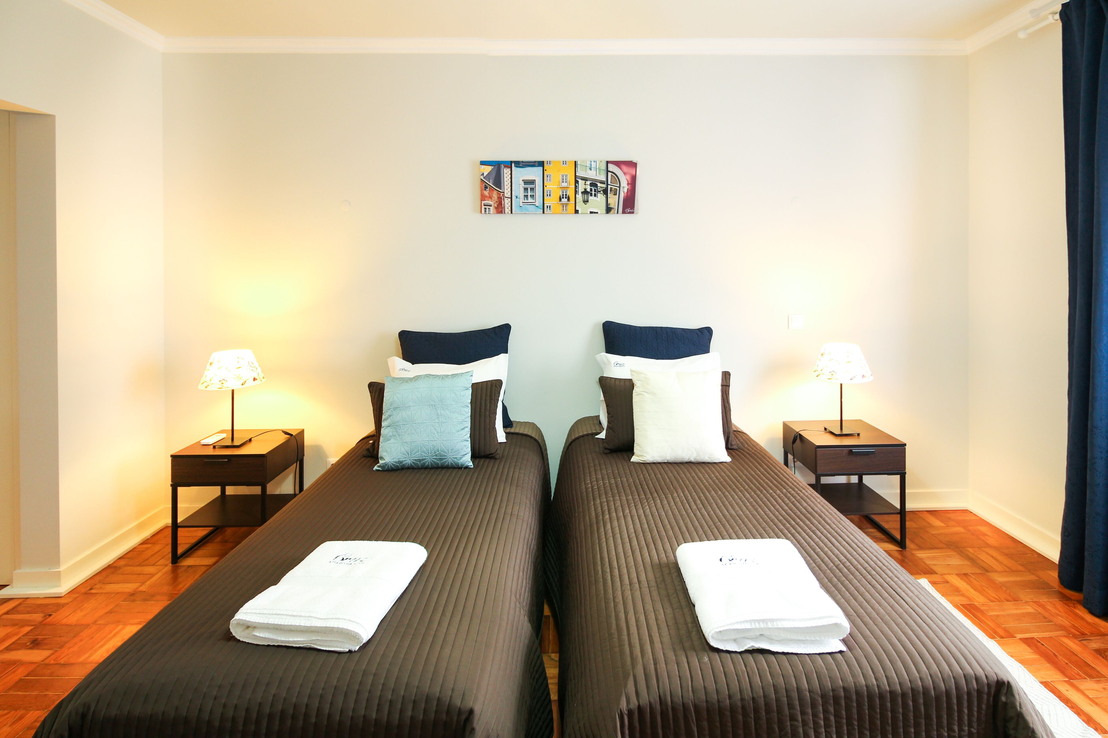 A hotel room with two beds, each with a pillow and a folded towel. There are bedside tables with lamps, a wall art piece above the beds, and a wooden floor.