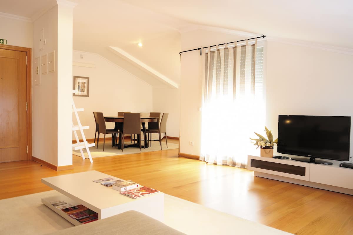 A modern living room with a flat-screen TV, wooden flooring, a dining area with chairs, and a staircase leading to another level.