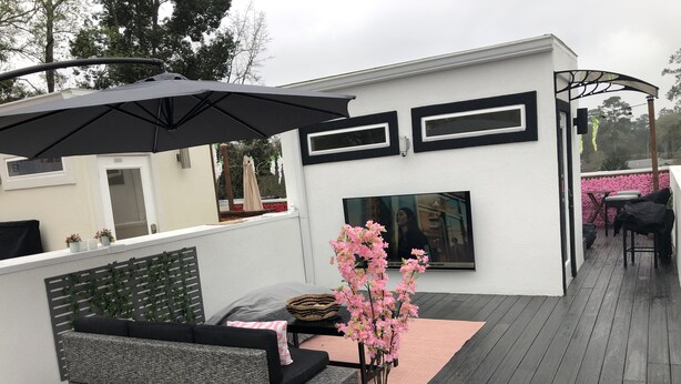 Zona d'estar exterior moderna amb un sofà gris, un para-sol negre i una planta en test amb flors roses.