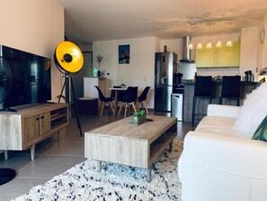 A modern living room with a TV, coffee table, and sofa. There is a dining area with chairs and a table. The kitchen is visible with cabinets and appliances.