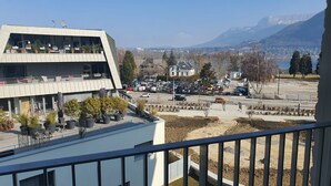 Un edificio moderno con tejado plano y varios balcones, rodeado de un estacionamiento y vistas a un paisaje montañoso.