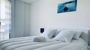 Una cama bien hecha con cabecera blanca y almohadas, una mesita de noche con lámpara, y una ventana con cortinas translúcidas.