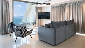 Een moderne woonkamer met een grijze bank, een stoel met zwart-wit patroon, een flatscreen televisie en een groot raam met uitzicht op zee.