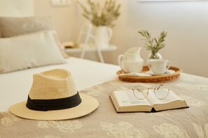 Una cama con una colcha color beige, un sombrero de paja con cinta negra, un libro abierto y un par de gafas sobre una bandeja con una planta pequeña.
