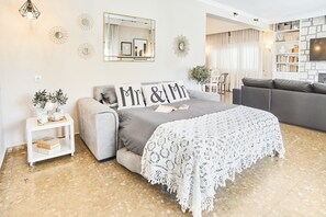 Un dormitorio con una cama, una mesita de noche blanca, un sofá gris, una mesa de centro blanca y una pared blanca decorada con espejos y obras de arte.