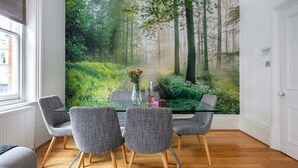 Un coin repas avec une table en verre et quatre chaises grises, une fresque représentant une forêt sur le mur et une fenêtre avec des stores.