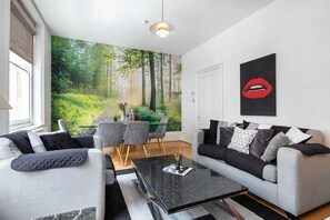 Uma sala de estar moderna com dois sofás cinza, uma mesa de centro preta e um mural com tema de floresta na parede.