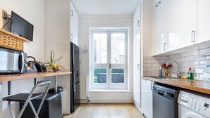 Une cuisine compacte équipée d’un four à micro-ondes, d’un grille-pain et d’un lave-vaisselle. Une fenêtre offre une vue sur les bâtiments environnants.