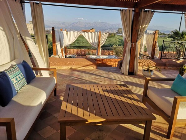 A terrace with a wooden table and two sofas, a hot tub, and a view of mountains.