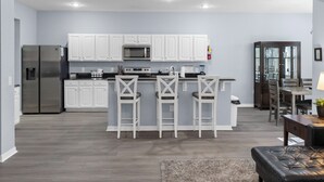 Une cuisine moderne avec des armoires blanches, un réfrigérateur en acier inoxydable, un four à micro-ondes et une cuisinière à gaz. On trouve trois tabourets de bar blancs autour de l’îlot central de la cuisine.