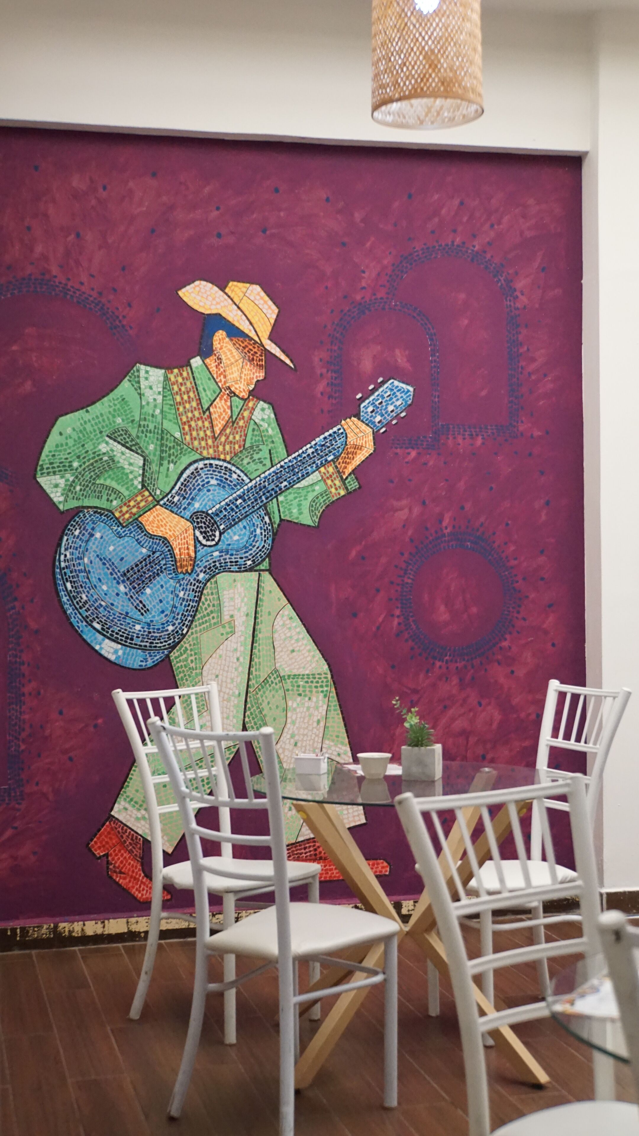 Une pièce décorée d’une grande fresque représentant une personne jouant de la guitare, avec des chaises blanches et une table sur laquelle est posée une petite plante.