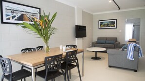 A modern living room with a dining area, a grey sofa, a TV mounted on the wall, and a large window. - Apartments at Mission Beach (Mission Beach)