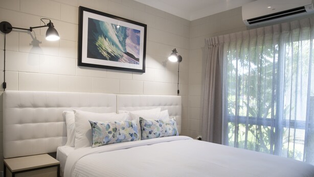 A bedroom with a white headboard, a framed picture on the wall, two bedside lamps, and a window with curtains. - Apartments at Mission Beach (Mission Beach)