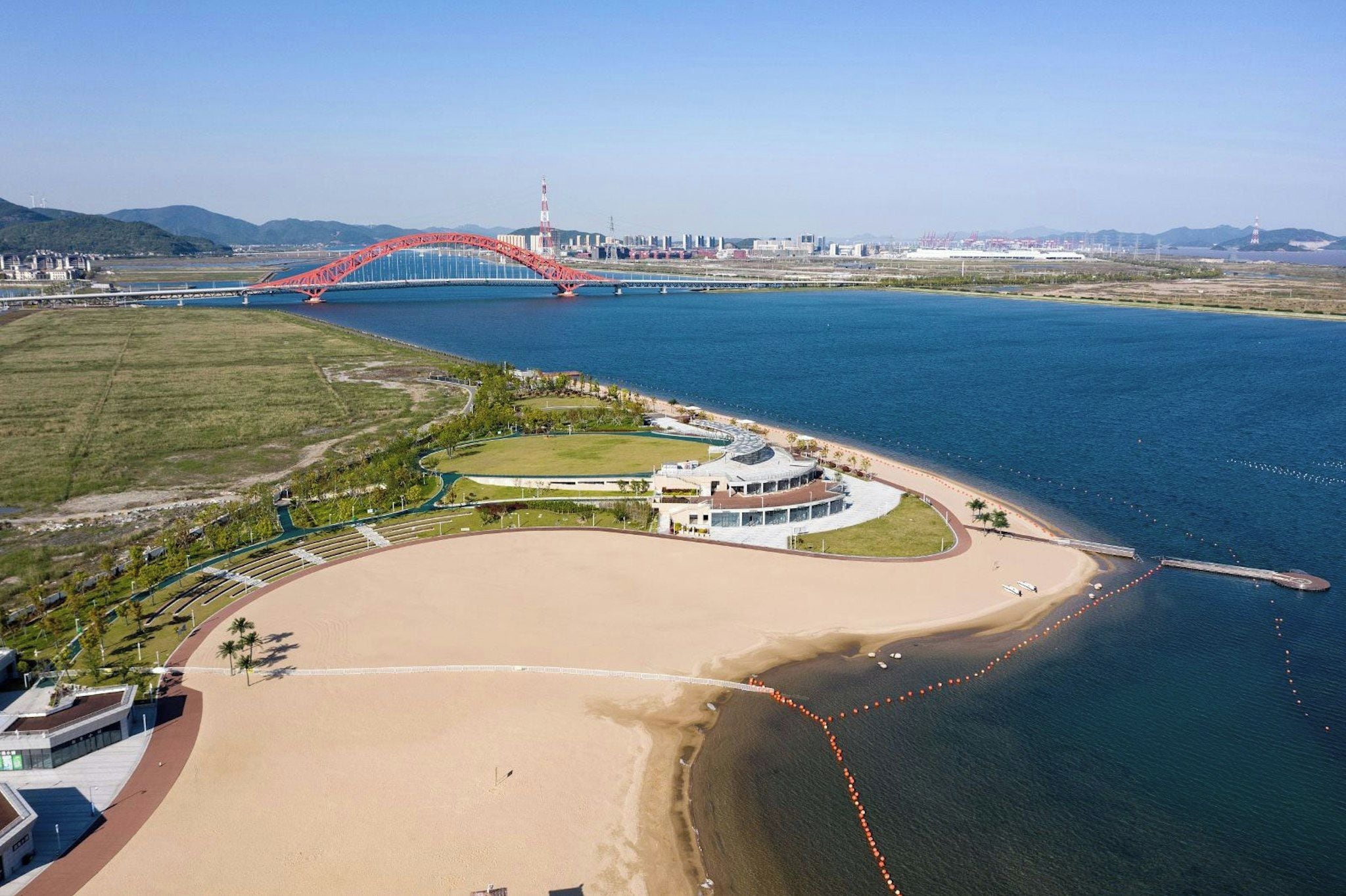 An aerial view of a coastal area with a large sandy beach, a marina, and a prominent red bridge in the background.