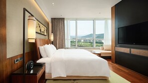 Une chambre d’hôtel avec un grand lit, une télévision, une table de chevet avec une lampe, une vue sur le paysage urbain à travers de grandes fenêtres et un plancher en bois.