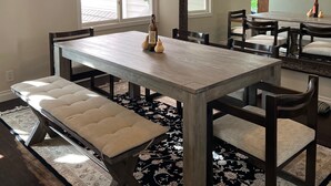 Une salle à manger avec une grande table, des bancs et des chaises. Un miroir est accroché au mur et un luminaire est fixé au plafond.