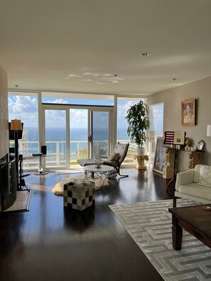 A spacious living room with a large window offering a view of the ocean. There is a comfortable seating area with a sofa and a chair, a coffee table, and a decorative plant. - "Cape Cod Beach-House" Panoramic Ocean View w/Hot Tub-5 min to Beach (Laguna Beach)